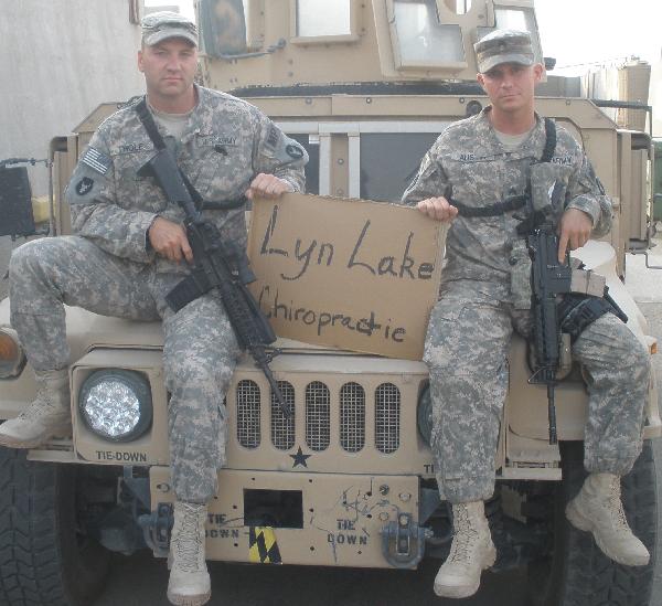 Two soldiers in camouflage uniforms seated on a military vehicle holding a handwritten Lyn Lake Chiropractic sign